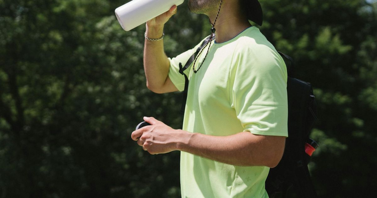 Sportivo uomo di profilo in mezzo alla natura, con maglietta verde, che beve da una borraccia. Foto per articolo sul tema Come fare allenamento in estate per non perdere i sali minerali?