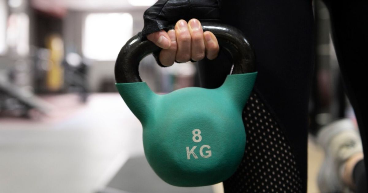 Dettaglio di una mano che indossa un guantino tecnico nero mentre solleva una kettlebell verde acqua da 8 kg in una palestra. Sullo sfondo sfocato si intravedono le attrezzature sportive.