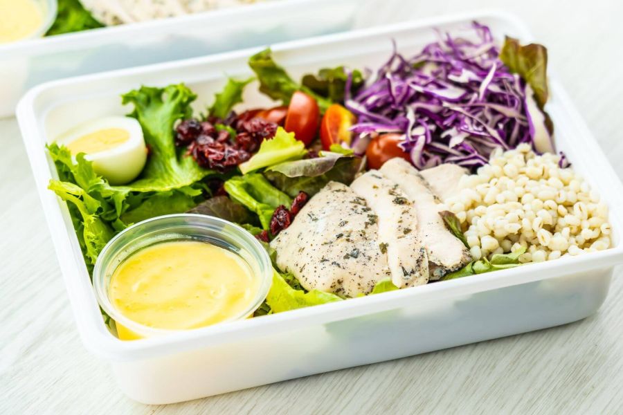 Piatto di insalata fresca con pollo grigliato, uovo sodo, orzo, pomodorini, insalata verde, cavolo rosso e salsa in un contenitore trasparente. Il piatto è ben equilibrato, con ingredienti freschi e colorati disposti in modo ordinato.