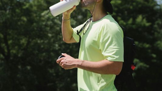 Sportivo uomo di profilo in mezzo alla natura, con maglietta verde, che beve da una borraccia. Foto per articolo sul tema Come fare allenamento in estate per non perdere i sali minerali?
