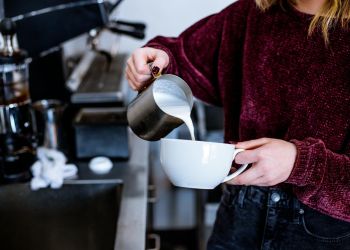 foto di una ragazza con un maglione che versa del latte da un bricco in una tazza per cappuccino, in cucina. foto per articolo su latte proteico