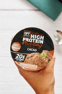 Mano che regge un vasetto di GFF High Protein Pudding al cacao su una tavola natalizia con ciotola di yogurt, noci e granella.
