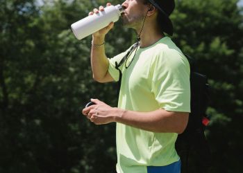 Sportivo uomo di profilo in mezzo alla natura, con maglietta verde, che beve da una borraccia. Foto per articolo sul tema Come fare allenamento in estate per non perdere i sali minerali?