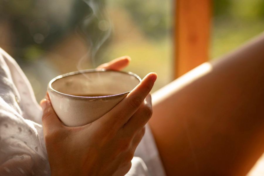 Primo piano sulle mani di una persona seduta in un ambiente caldo e luminoso, che tiene una tazza di bevanda calda da cui sale del vapore. La scena evoca un momento di relax, con la luce dorata del tardo pomeriggio o della sera che illumina parte della gamba e una camicia bianca.