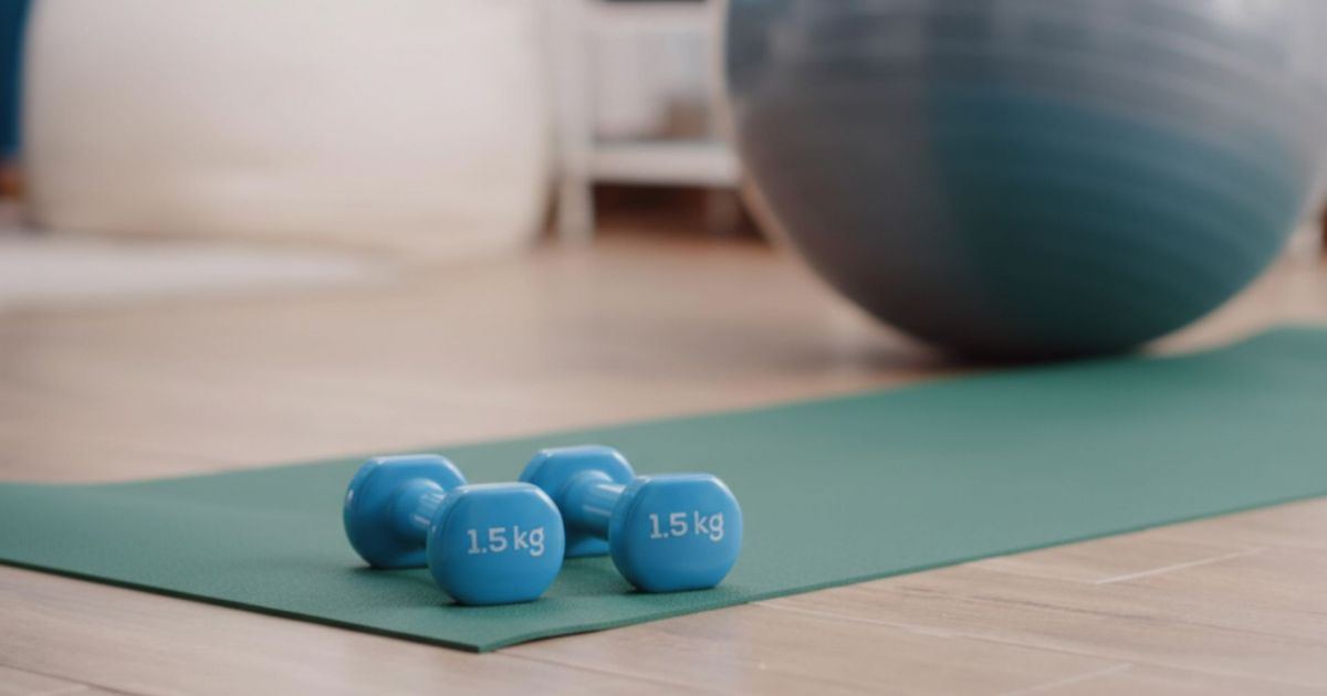Inquadratura ravvicinata ad angolo basso di un tappetino da fitness verde disteso su un pavimento in legno. Sopra il tappetino sono appoggiati due manubri azzurri da 1,5 kg. Sullo sfondo sfocato si vedono una fitball grigia e un ambiente domestico luminoso