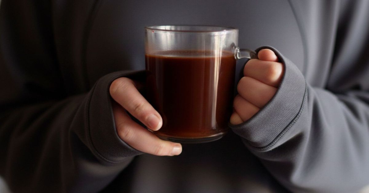 Primo piano delle mani di una ragazza che regge una tazza di vetro trasparente colma di cioccolata calda densa e scura, indossando una felpa tecnica grigio antracite