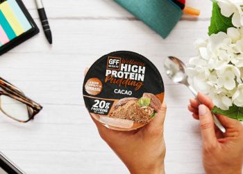 Mano che regge un vasetto di GFF High Protein Pudding al cacao su una tavola natalizia con ciotola di yogurt, noci e granella.