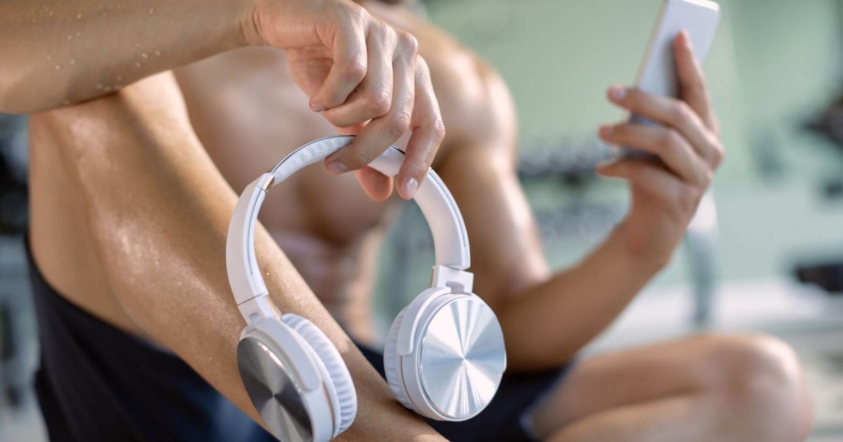 Persona seduta in palestra, con indosso un outfit sportivo, che tiene in mano delle cuffie bianche e uno smartphone. L'immagine suggerisce un momento di pausa durante l'allenamento, con una leggera luminescenza sulla pelle, evidenziando l'energia e la concentrazione