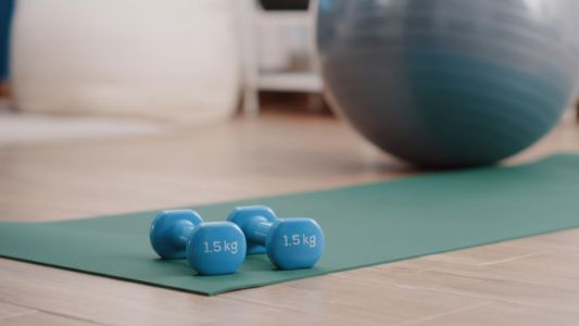 Inquadratura ravvicinata ad angolo basso di un tappetino da fitness verde disteso su un pavimento in legno. Sopra il tappetino sono appoggiati due manubri azzurri da 1,5 kg. Sullo sfondo sfocato si vedono una fitball grigia e un ambiente domestico luminoso