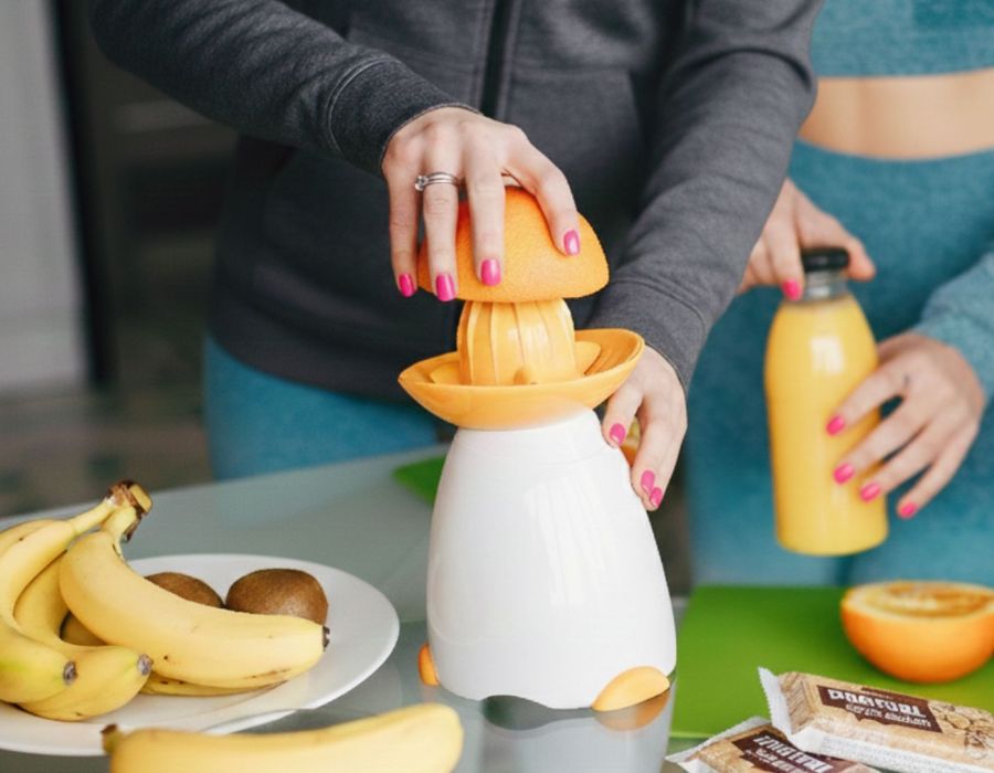 Due donne in abbigliamento sportivo, con felpa grigia e verde acqua. Una tiene un preparato di succo d'arancia e un'arancia intera in una cucina moderna e luminosa.