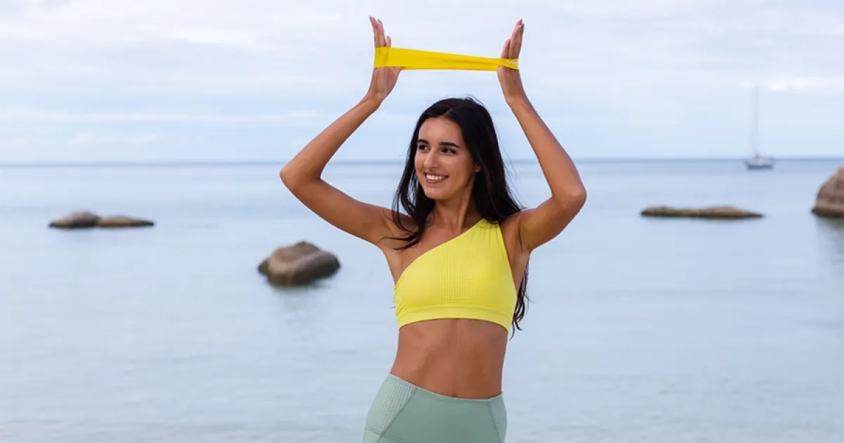 Sportiva sorridente che esegue un esercizio con un elastico in spiaggia, un'idea di workout per la routine estiva GFF.