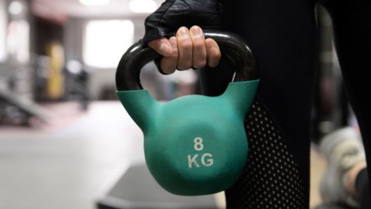 Dettaglio di una mano che indossa un guantino tecnico nero mentre solleva una kettlebell verde acqua da 8 kg in una palestra. Sullo sfondo sfocato si intravedono le attrezzature sportive.