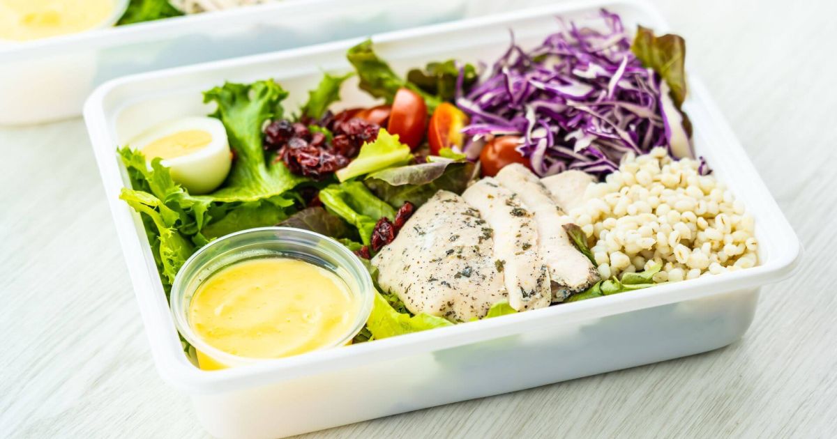 Piatto di insalata fresca con pollo grigliato, uovo sodo, orzo, pomodorini, insalata verde, cavolo rosso e salsa in un contenitore trasparente. Il piatto è ben equilibrato, con ingredienti freschi e colorati disposti in modo ordinato.