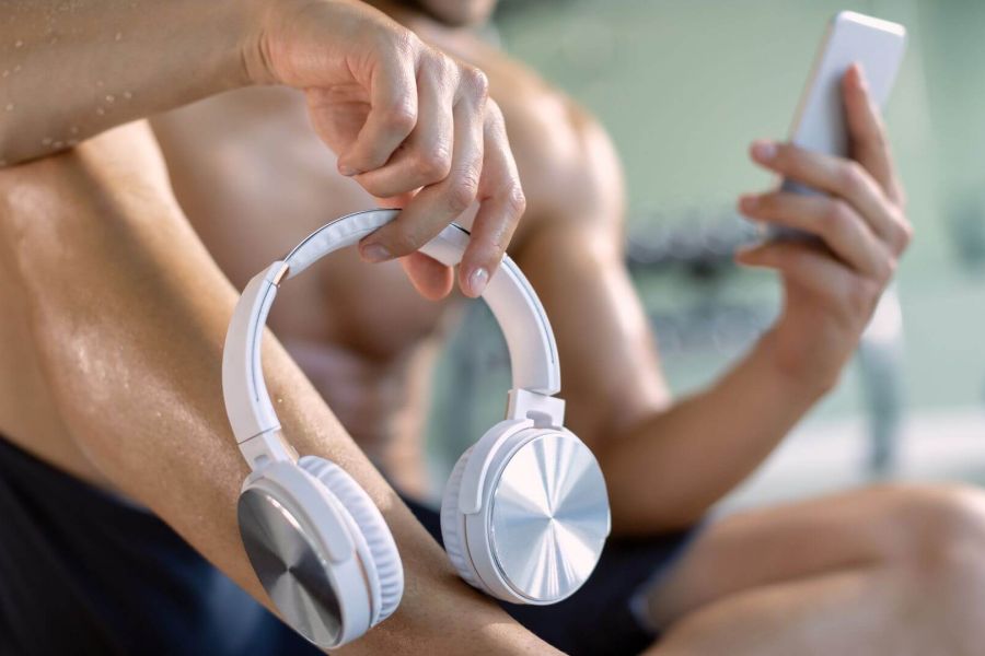 Persona seduta in palestra, con indosso un outfit sportivo, che tiene in mano delle cuffie bianche e uno smartphone. L'immagine suggerisce un momento di pausa durante l'allenamento, con una leggera luminescenza sulla pelle, evidenziando l'energia e la concentrazione
