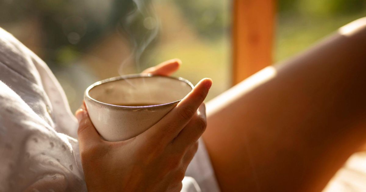 Primo piano sulle mani di una persona seduta in un ambiente caldo e luminoso, che tiene una tazza di bevanda calda da cui sale del vapore. La scena evoca un momento di relax, con la luce dorata del tardo pomeriggio o della sera che illumina parte della gamba e una camicia bianca.