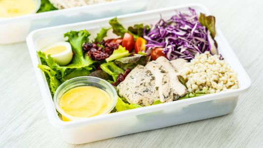 Piatto di insalata fresca con pollo grigliato, uovo sodo, orzo, pomodorini, insalata verde, cavolo rosso e salsa in un contenitore trasparente. Il piatto è ben equilibrato, con ingredienti freschi e colorati disposti in modo ordinato.