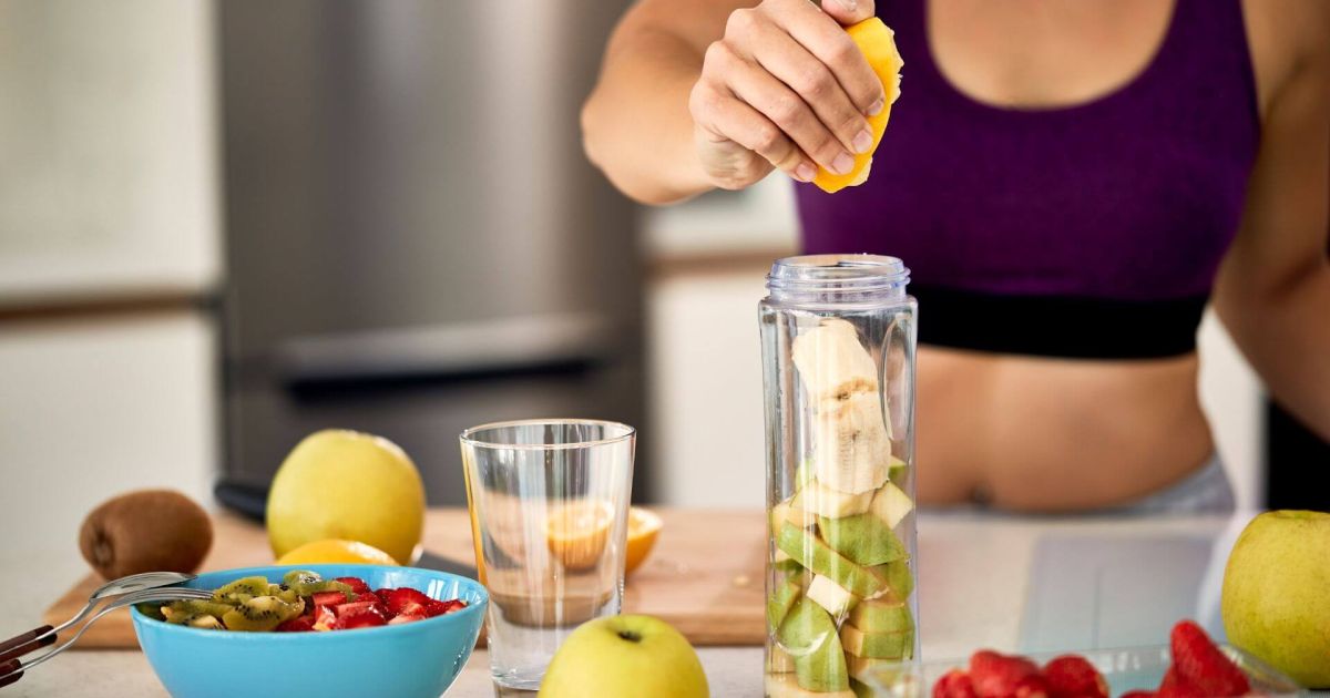 Una persona con un top sportivo viola sta spremendo un pezzo di agrume in un frullatore che contiene pezzi di banana e mela verde. Sul bancone ci sono una ciotola con fragole e kiwi tagliati, un bicchiere vuoto e altri frutti interi come mele e kiwi. L'immagine suggerisce la preparazione di un frullato o uno spuntino salutare.