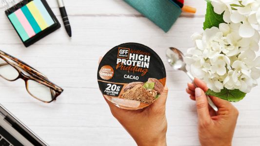 Mano che regge un vasetto di GFF High Protein Pudding al cacao su una tavola natalizia con ciotola di yogurt, noci e granella.