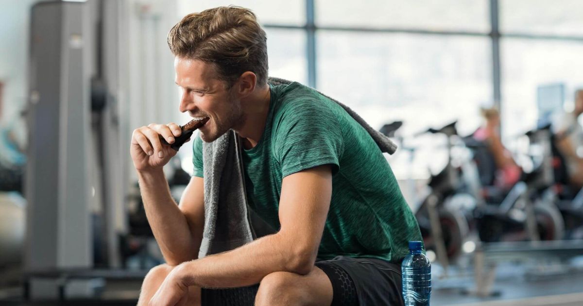 Uomo seduto in palestra, con una maglia verde e un asciugamano sulle spalle, che sta mangiando una barretta proteica mentre tiene in mano una bottiglia d'acqua. Sullo sfondo si vedono attrezzature da palestra e altre persone che si allenano.