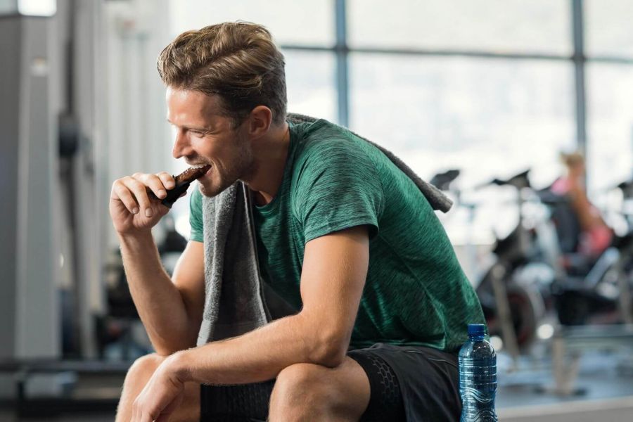 Uomo seduto in palestra, con una maglia verde e un asciugamano sulle spalle, che sta mangiando una barretta proteica mentre tiene in mano una bottiglia d'acqua. Sullo sfondo si vedono attrezzature da palestra e altre persone che si allenano.