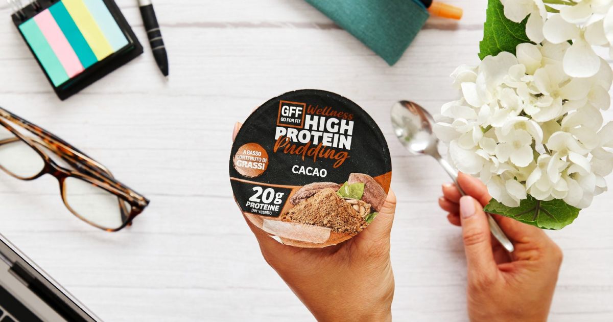 Mano che regge un vasetto di GFF High Protein Pudding al cacao su una tavola natalizia con ciotola di yogurt, noci e granella.