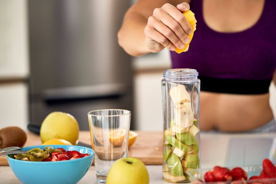Una persona con un top sportivo viola sta spremendo un pezzo di agrume in un frullatore che contiene pezzi di banana e mela verde. Sul bancone ci sono una ciotola con fragole e kiwi tagliati, un bicchiere vuoto e altri frutti interi come mele e kiwi. L'immagine suggerisce la preparazione di un frullato o uno spuntino salutare.